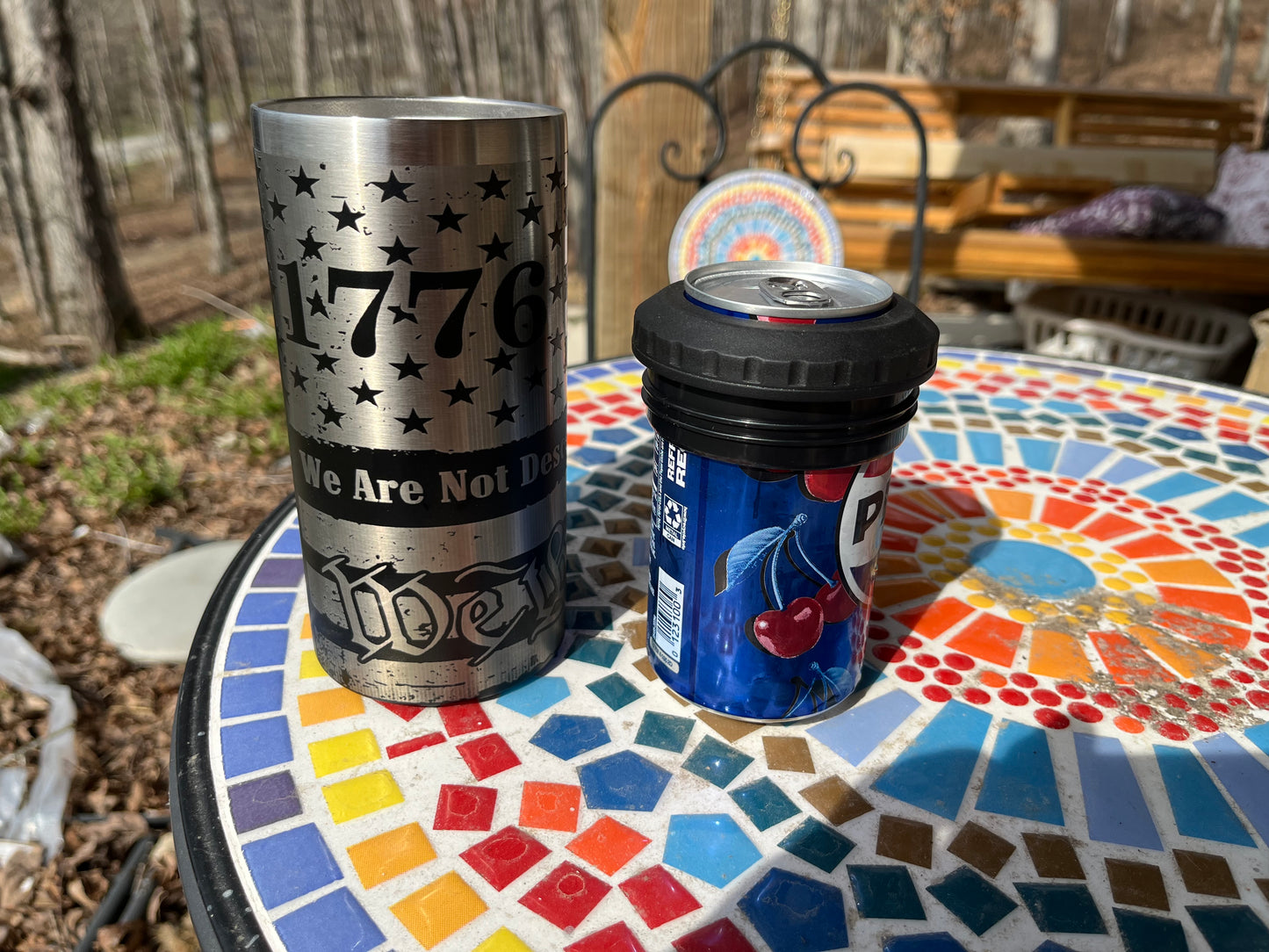 Metal tumbler with '1776 We Are Not Divided' text and a can of soda on a mosaic table outdoors.