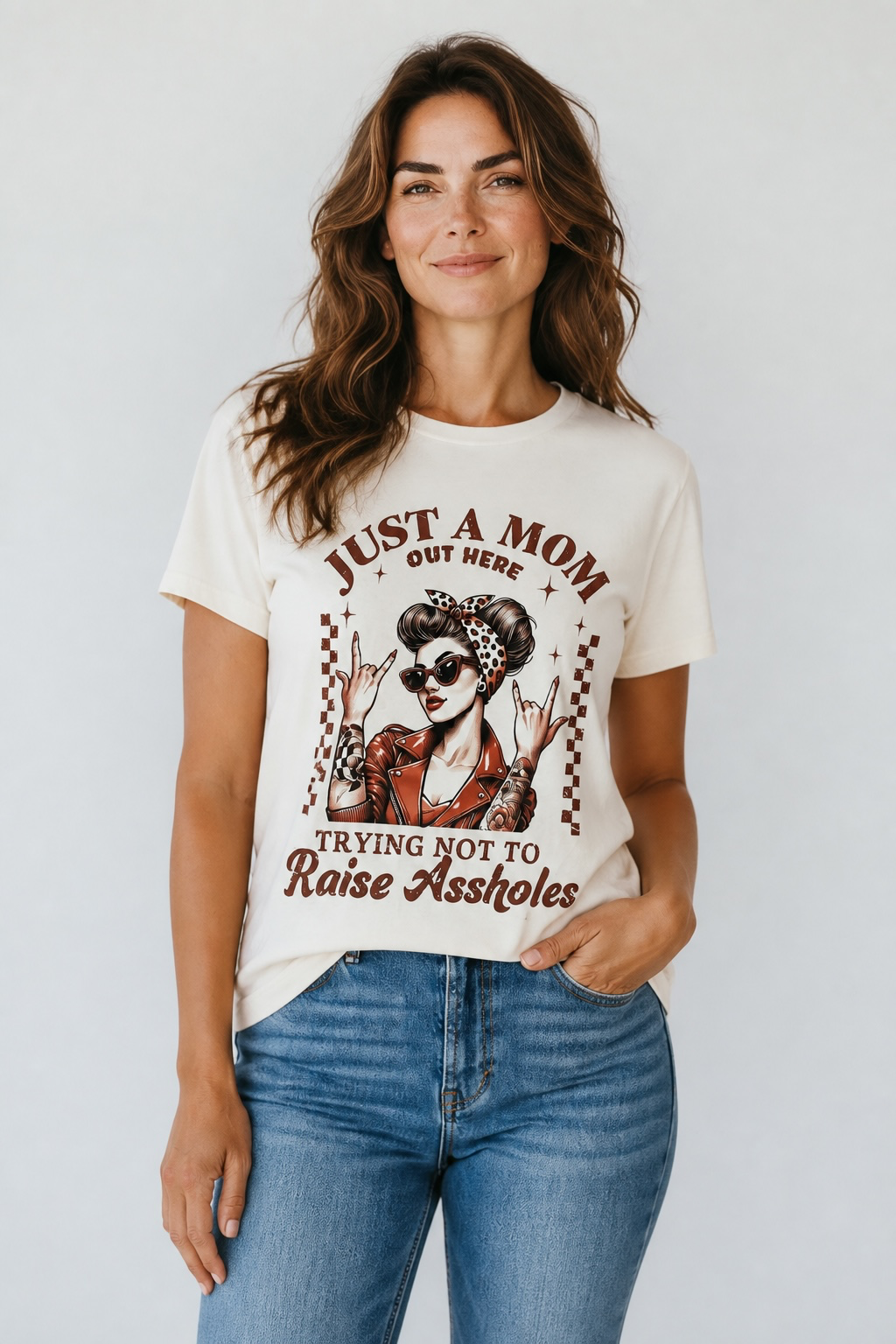 A woman wearing a cream-colored graphic t-shirt with a retro-style biker mom illustration and the text 'JUST A MOM OUT HERE TRYING NOT TO Raise Assholes', paired with blue jeans, against a light gray studio background.