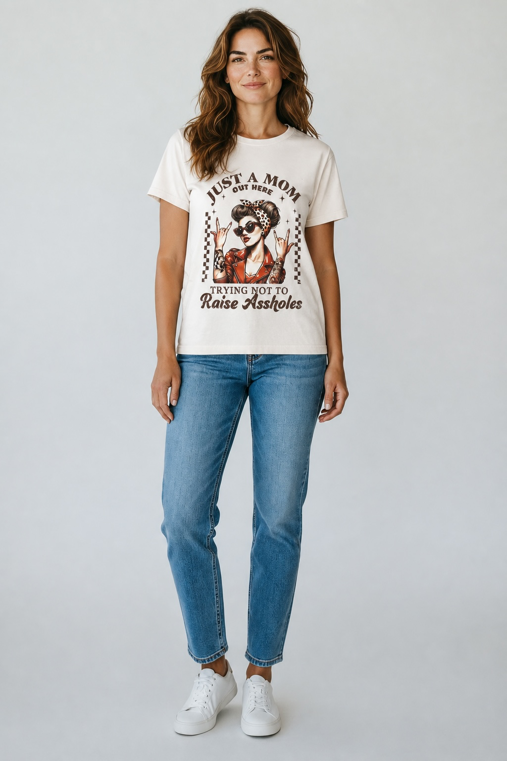 A woman modeling a cream-colored graphic t-shirt with a distressed design featuring a dog and the text 'JUST A MOM TRYING NOT TO Raise Assholes', paired with blue jeans and white sneakers, against a light gray studio background.