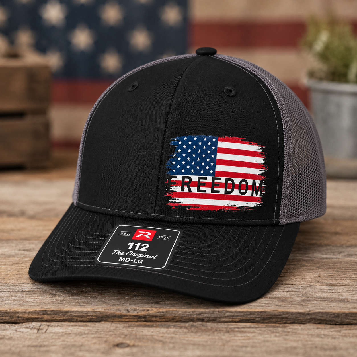 A black and gray trucker cap with a distressed American flag graphic and the word 'FREEDOM' on the front, displayed on a rustic wooden surface with an American flag and potted plants blurred in the background.